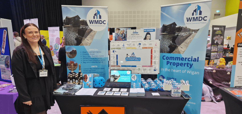 Image shows female business owner smiling standing beside her exhibition stand