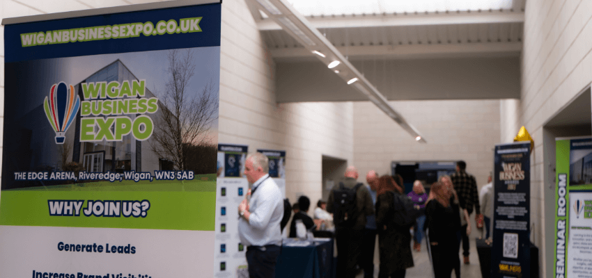 A convention hall with a sign that reads Wigan Business Expo and a crowd of people networking in the background.
