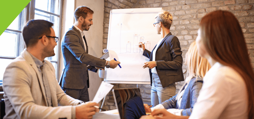 Business workshop meeting with a presenter explaining a plan on a flipchart while attendees listen and take notes.