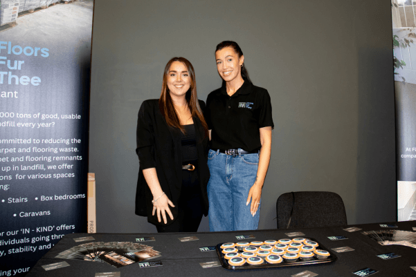 Two exhibitors smiling behind their stand at Wigan Business Expo, with promotional materials displayed on the table.